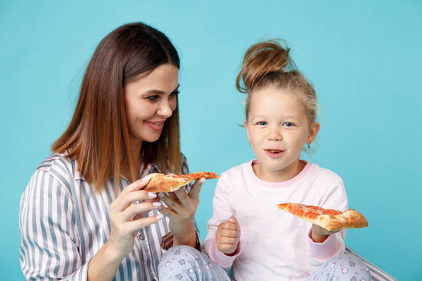 Mutter und Tochter beim Pizza essen auf dem Boden sitzend im Schlafanzug vor blauem Hintergrund. - Foto, Bild