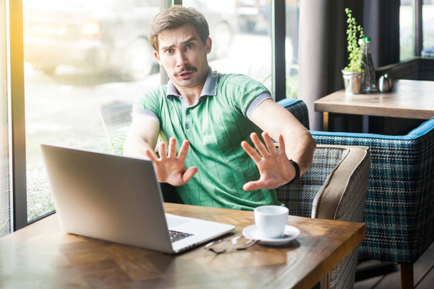 Young scared businessman in green t-shirt looking with afraid face and showing at camera stop sign while sitting at table with laptop in cafe, business and freelancing concept  - 写真・画像