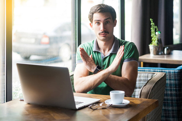 Young serious businessman in green t-shirt looking at camera and showing closed sign with hands while sitting at table with laptop in cafe, business and freelancing concept - Φωτογραφία, εικόνα