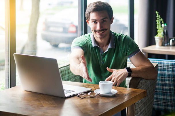 Young happy businessman in green t-shirt looking at camera and giving hand to greeting while sitting at table with laptop in cafe, business and freelancing concept - Foto, immagini
