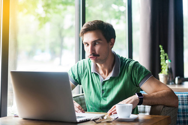 Young funny businessman in green t-shirt looking at laptop display with stupid crazy face and crossed eyes while sitting at table in cafe, business and freelancing concept - Zdjęcie, obraz