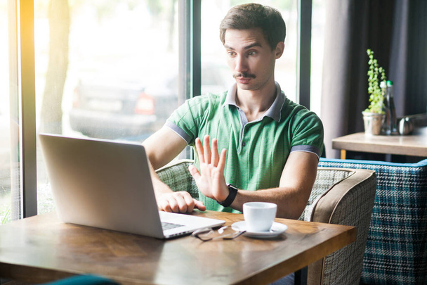 Young serious businessman in green t-shirt looking at laptop screen and showing stop sign gesture while sitting at table in cafe, business and freelancing concept  - Zdjęcie, obraz