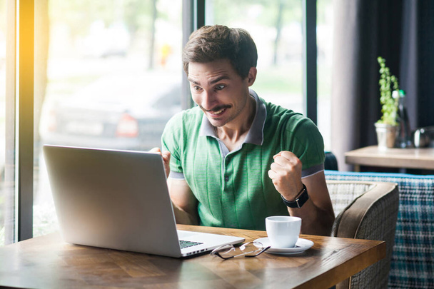 Young happy surprised businessman in green t-shirt looking at laptop screen and rejoicing success while sitting at table in cafe, business and freelancing concept  - Foto, afbeelding