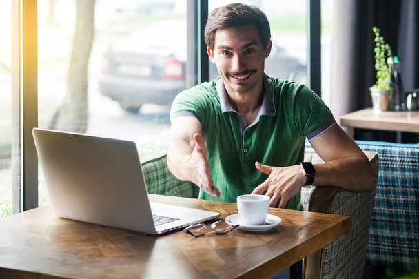 Young happy businessman in green t-shirt looking at camera and giving hand to greeting while sitting at table with laptop in cafe, business and freelancing concept - Fotó, kép
