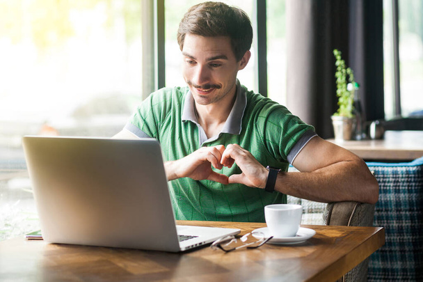 Young happy romantic businessman in green t-shirt looking at laptop screen during video call and showing love gesture on heart  while sitting at table in cafe, business and freelancing concept  - Photo, Image