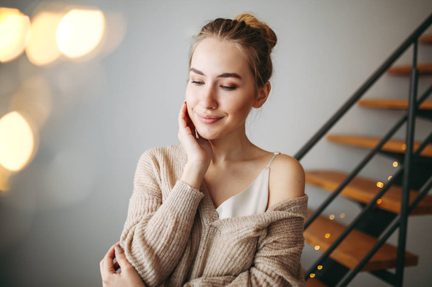 Młoda piękna dziewczyna marzy z blond włosami wieczorem beżowy jedwabny strój i przytulny sweter przed schodami w domu - Zdjęcie, obraz