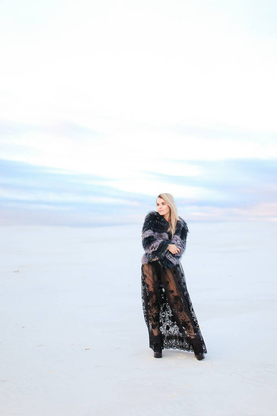 Joven mujer de moda con vestido negro de pie en el fondo de la estepa de invierno blanco
. - Foto, imagen