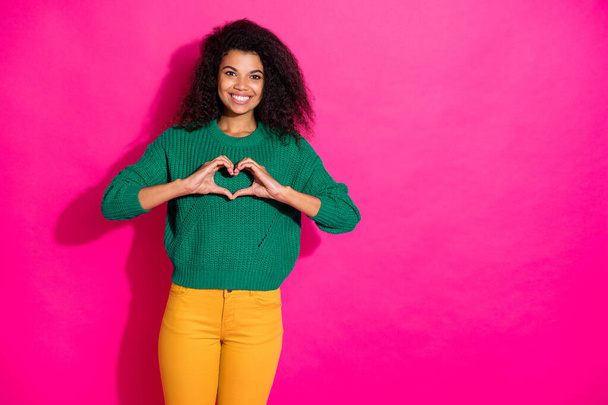 Portrait of positive dreamy afro american girl show heart from fingers symbol of her love to boyfriend on valentine day in winter wear knitted pullover yellow pants isolated pink color background - Valokuva, kuva