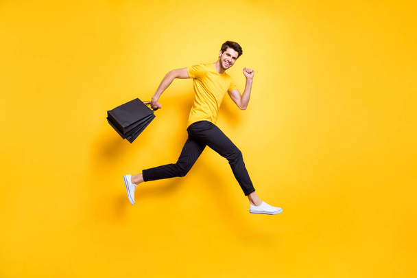 Full size photo of handsome guy jumping high holding many packages speed rushing sale shopping wear casual t-shirt black pants isolated yellow color background - Фото, зображення