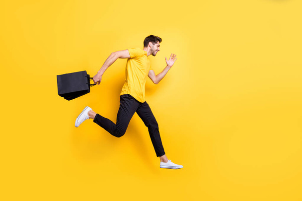 Full size photo of handsome guy jumping high holding packages speed rushing sale shopping wear casual t-shirt pants isolated yellow color background - Fotoğraf, Görsel
