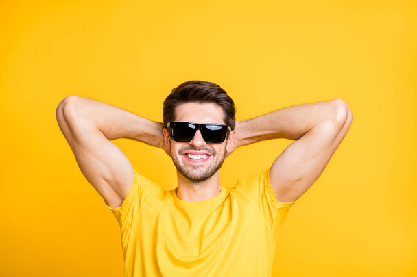 Photo of handsome carefree guy relaxing summer vacation abroad holding hands behind head sun bathing wear sun specs casual t-shirt isolated yellow color background - Foto, afbeelding