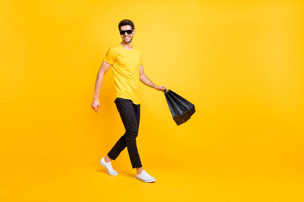 Full body photo of handsome guy carry boutique bags make abroad shopping go fashionable mall wear casual t-shirt black pants isolated yellow color background - Fotoğraf, Görsel