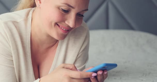 Adult  woman using mobile phone, at home. Adult beautiful  woman typing message  on a smartphone. Happy smiling woman uses cell phone lying on bed. Cheerful woman holds  smart phone.         - Materiał filmowy, wideo