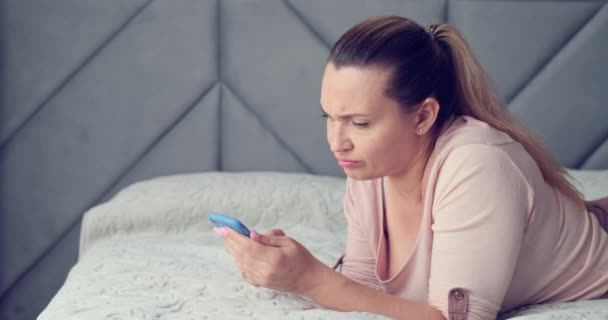 Adult woman upset by the bad news. Beautiful adult white woman is chatting on the smart phone. Woman is shocked by a bad sms message. Caucasian female lying on a bed with cell phone. Real time - Footage, Video