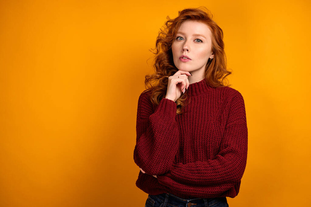 Red-haired girl in a burgundy sweater is standing on an orange background and propping chin with her hand, looks into the frame - Photo, image