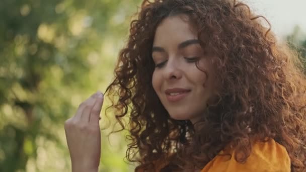Pretty lovely young redhead woman in orange dress smiling and playing with her curly hair while resting in the park - 映像、動画