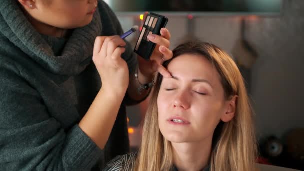 Make-up artist applying bright base color eyeshadow on models eye and holding a shell with eyeshadow on background, close up - Záběry, video
