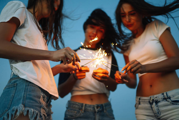 Young happy girls stands on beach with sparkler in sunset light. Teenage girls with sparklers celebrate and laugh. Party, holidays, nightlife and people concept. - Foto, immagini