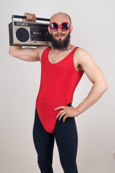 Portrait of a funny bearded man in a red bodysuit and black leggings holding a retro tape recorder on a white background - Fotoğraf, Görsel