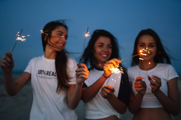 Young happy girls stands on beach with sparkler in sunset light. Teenage girls with sparklers celebrate and laugh. Party, holidays, nightlife and people concept. - 写真・画像