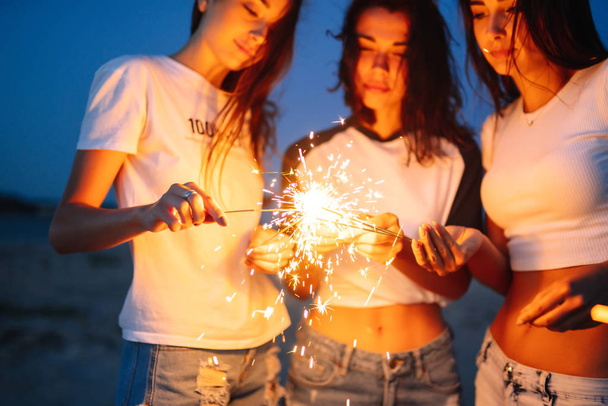 Young happy girls stands on beach with sparkler in sunset light. Teenage girls with sparklers celebrate and laugh. Party, holidays, nightlife and people concept. - Zdjęcie, obraz