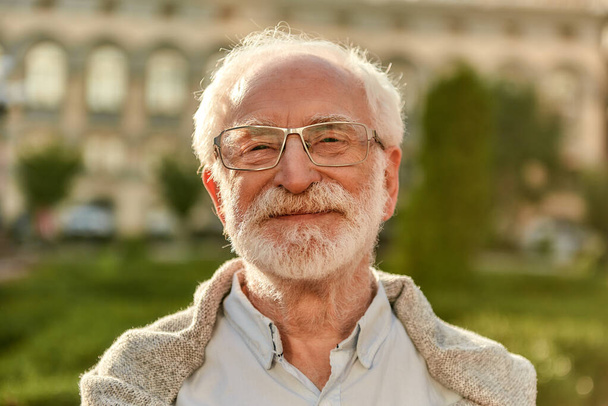 Θετική στάση. Πορτρέτο του όμορφου γενειοφόρου ηλικιωμένου με γυαλιά κοιτάζοντας την κάμερα και χαμογελώντας ενώ περνούν το χρόνο τους σε εξωτερικούς χώρους σε μια ηλιόλουστη μέρα - Φωτογραφία, εικόνα