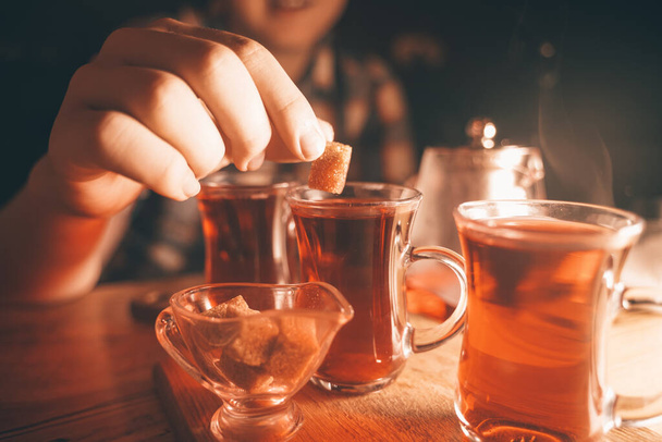 Ένας νεαρός έφηβος βάζει κίτρινη ζάχαρη σε ένα φλιτζάνι τσάι, κρατώντας την στα χέρια του πάνω από ένα ποτήρι. - Φωτογραφία, εικόνα
