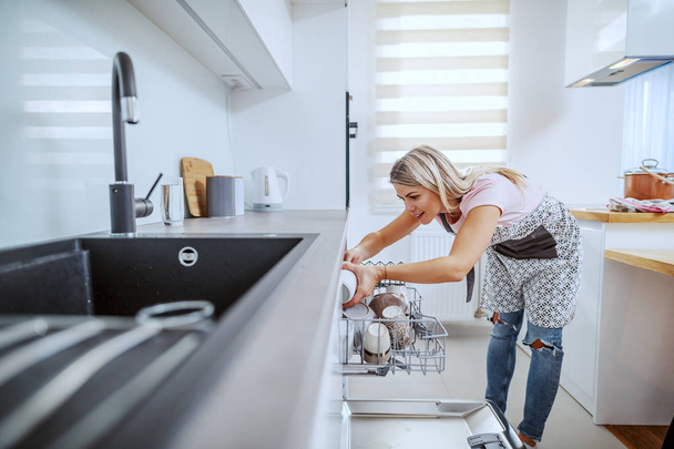 Digne caucasienne blonde femme au foyer dans tablier debout dans la cuisine domestique et mettre la vaisselle au lave-vaisselle
. - Photo, image
