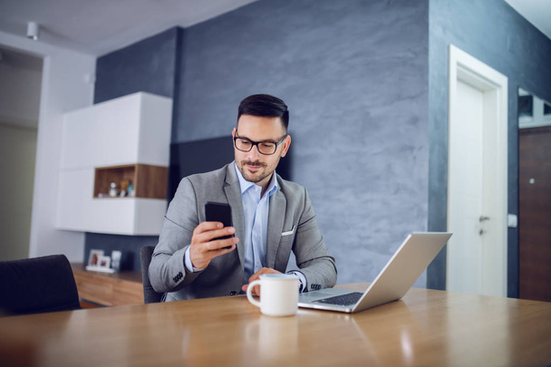 Charming caucasian bearded businessman with eyeglasses and in suit sitting at home and using smart phone. On table is laptop and mug with fresh morning coffee. - Photo, image