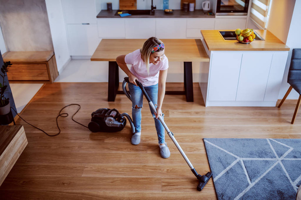 Top view of cheerful caucasian blond young housewife dressed casual using vacuum cleaner to clean floor in living room. - Foto, Imagem