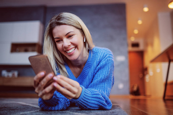 Charmante blanke blonde glimlachende vrouw in trui liggend op de buik op de vloer in de woonkamer en met behulp van een smartphone. In de oren zijn bluetooth oortelefoons. - Foto, afbeelding