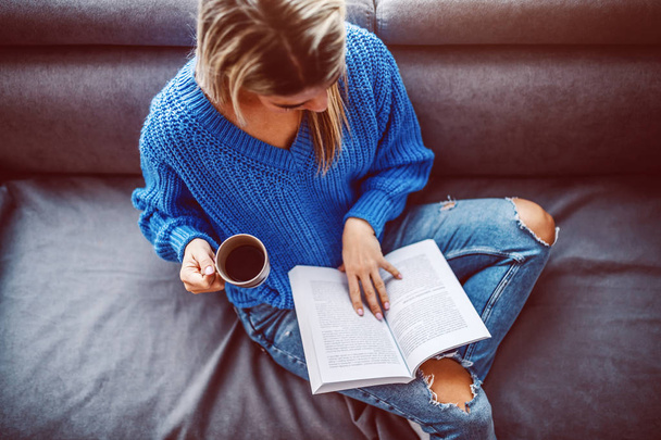 Top view of blond caucasian young woman in sweater sitting on sofa in living room, drinking coffee and reading book. - Foto, imagen