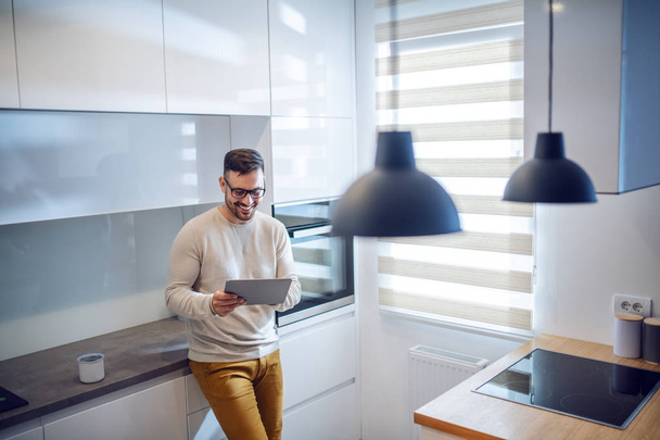 Handsome smiling caucasian unshaven man dressed casual and with eyeglasses leaning on kitchen counter and using tablet. - Foto, Imagen