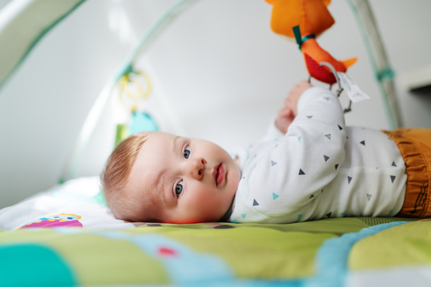 Seitenansicht des schönen neugierigen kaukasischen sechs Monate alten Jungen, der im Bett liegt und mit Krippenspielzeug spielt, während er in die Kamera schaut. - Foto, Bild