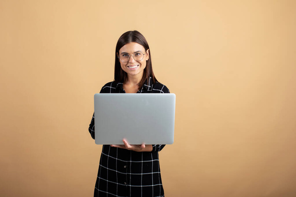 Een jonge vrouw in een geruite jurk staat op een oranje achtergrond met een laptop - Foto, afbeelding