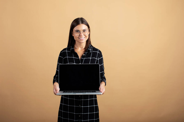 A young woman in a plaid dress stands on an orange background with a laptop - Foto, Imagen