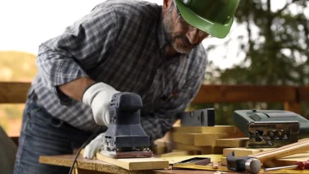 Adult carpenter craftsman wearing helmet and leather protective gloves, with the electric sander smoothes a wooden table. Construction industry, housework do it yourself. Safety at work. - Materiał filmowy, wideo
