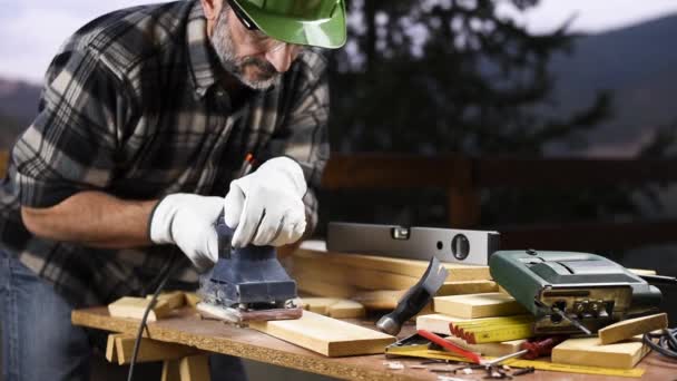Adult carpenter craftsman wearing helmet and leather protective gloves, with the electric sander smoothes a wooden table. Construction industry, housework do it yourself. Safety at work. - Πλάνα, βίντεο