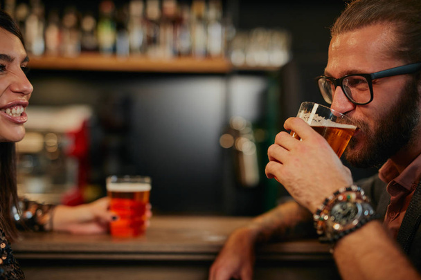 Young handsome caucasian bearded man with eyeglasses leaning on bar counter in pub, drinking beer and chatting with friend. - 写真・画像