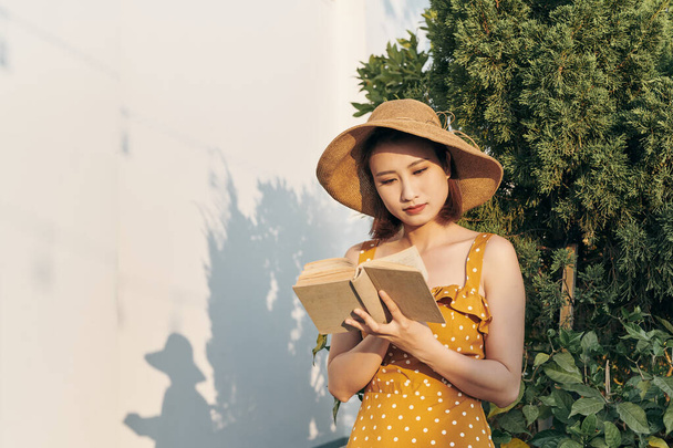 Mujer joven leyendo un libro de pie apoyado contra el árbol del tronco en el parque de verano al aire libre
. - Foto, imagen