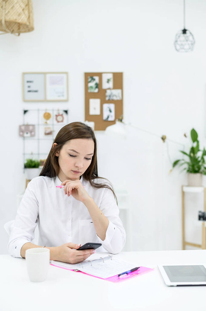 A young woman in a white shirt sits at a desk with a phone in her hands and plans a schedule. Female office worker makes a pen in the organizer. Paper notebook for notes. - Photo, image