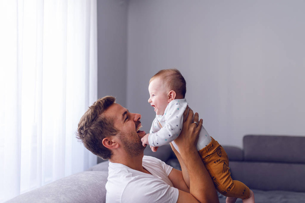 Zabawny przystojny biały młody tata podnosząc jego kochający 6 miesięcy chłopiec podczas siedzenia na kanapie w salonie. Dziecko cieszące się czasem z ojcem i śmiechem. - Zdjęcie, obraz