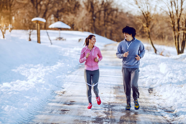 Feliz casal caucasiano sorridente em roupas esportivas correndo na natureza enquanto olham um para o outro. Hora de Inverno
. - Foto, Imagem