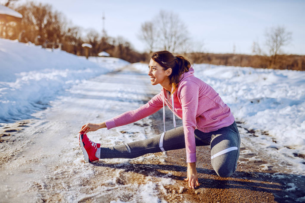 Side view of beautiful caucasian woman in sportswear and with ponytail stretching leg on country road. Winter time. Outdoor fitness concept. - Photo, Image