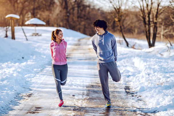fröhliches niedliches kaukasisches Paar in Sportkleidung, das sich auf der Landstraße die Beine ausstreckt und einander anschaut. Winterzeit. - Foto, Bild