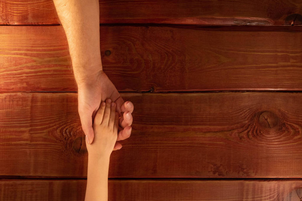 Children's and men's hands. Son placed his hand on his father's palm on background of red textured wooden table. Concept of family relations, help, kindness, understanding and love. Flat lay. - Photo, Image