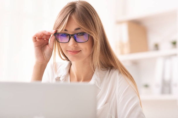 Lächelnde Dame blickt durch eine Brille auf Laptop im Büro - Foto, Bild