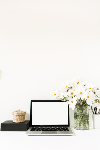Business, freelance, blogger concept. Mock up, empty, copy space. Laptop standing on white table against white wall with daisies in vase, black box, straw basket.  - Photo, Image