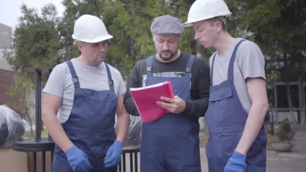 Two adult Caucasian movers in helmets and blue uniform standing with their chief. Three workers reading waybill or consignment, planning work. - Materiał filmowy, wideo