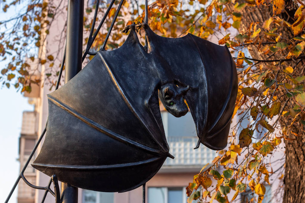 The black metal statue of a spooky bat handing on the tree in the city street park. - Photo, Image
